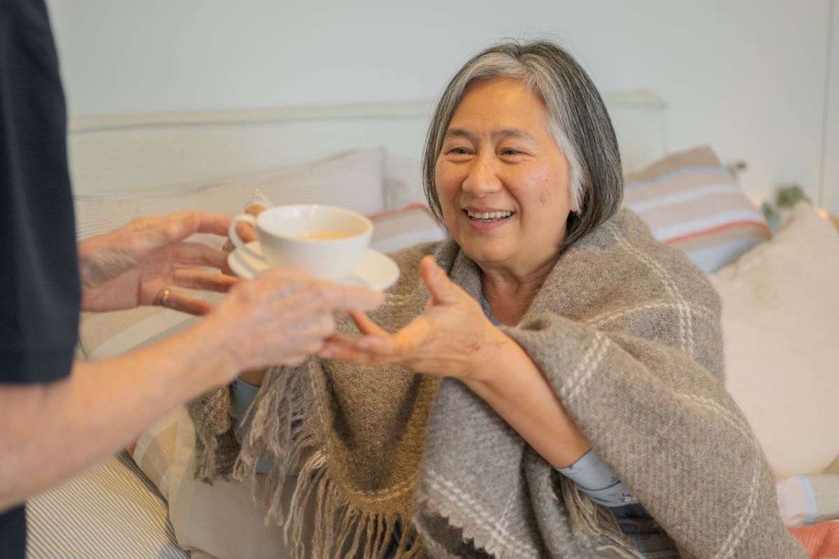 An elderly woman receives a cup of tea from a support worker.
