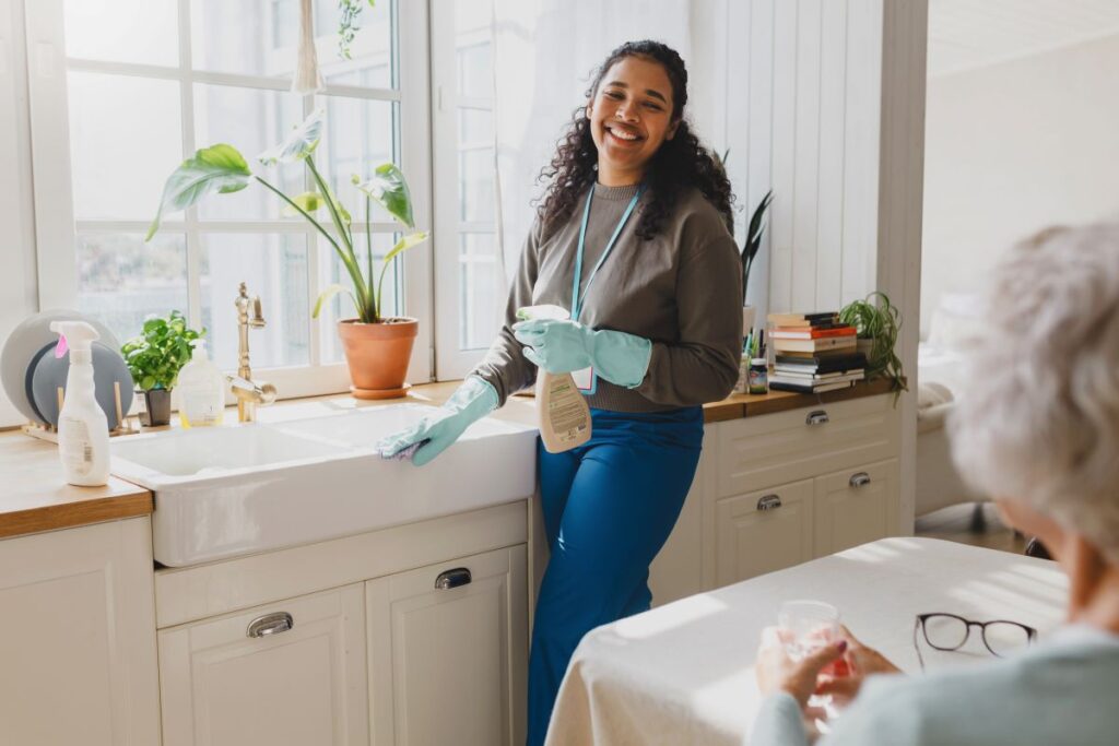 A support worker cleans the kitchen while an elderly woman watches.